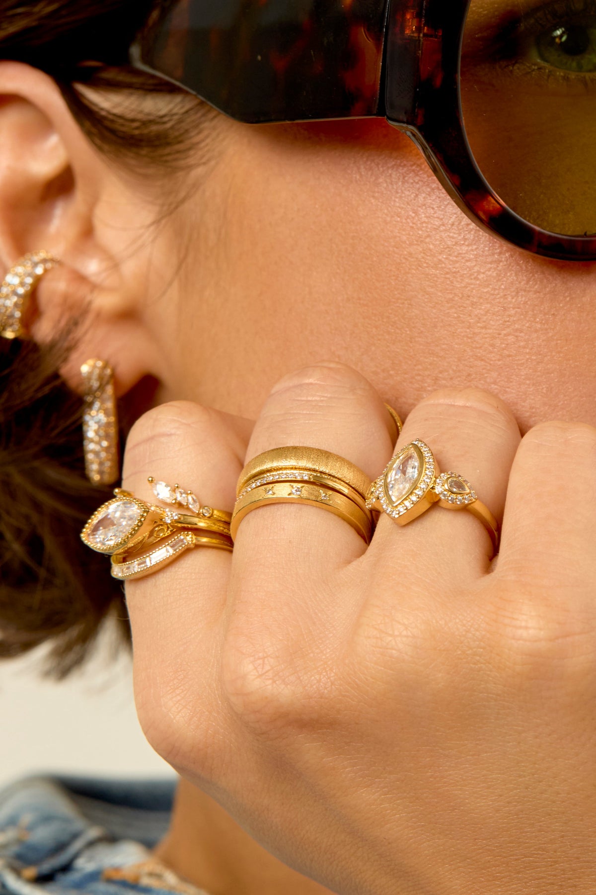 Model wearing multiple gold rings with crystal stones and hoop earrings, styled with sunglasses