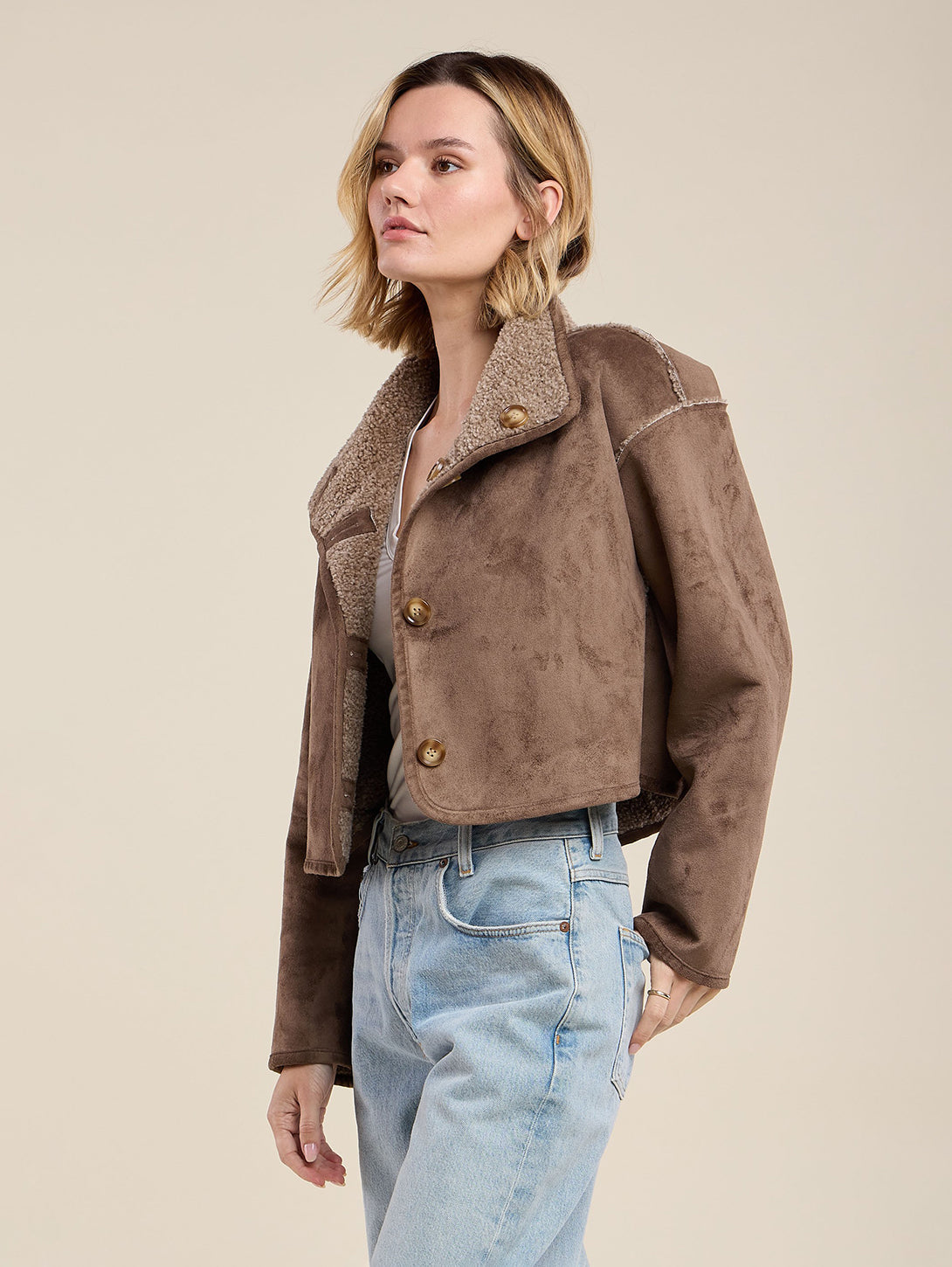Model wearing cropped brown faux shearling jacket with sherpa collar and gold buttons over light jeans