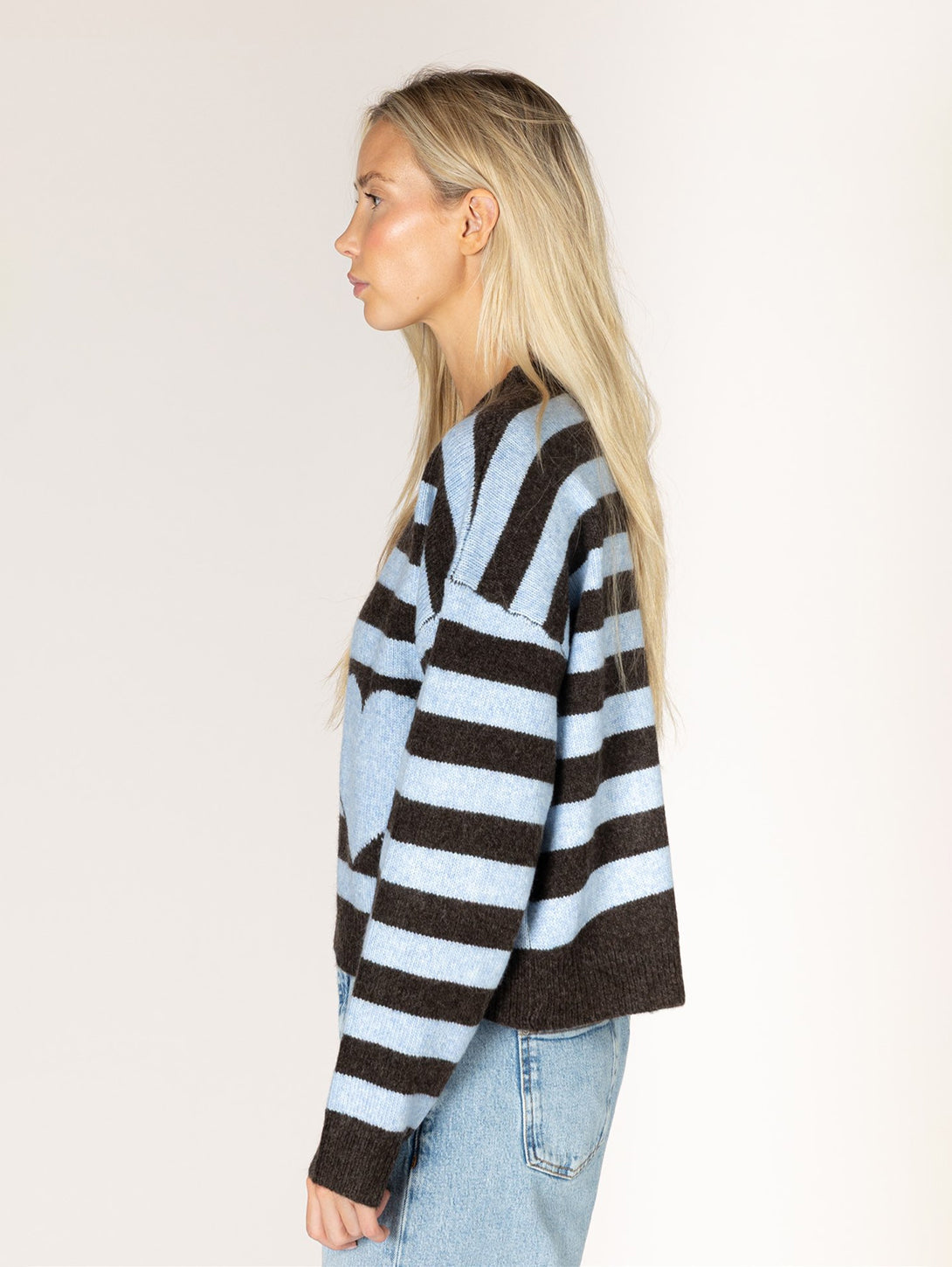 Side view of woman wearing brown and sky blue striped heart-detail knit sweater with jeans