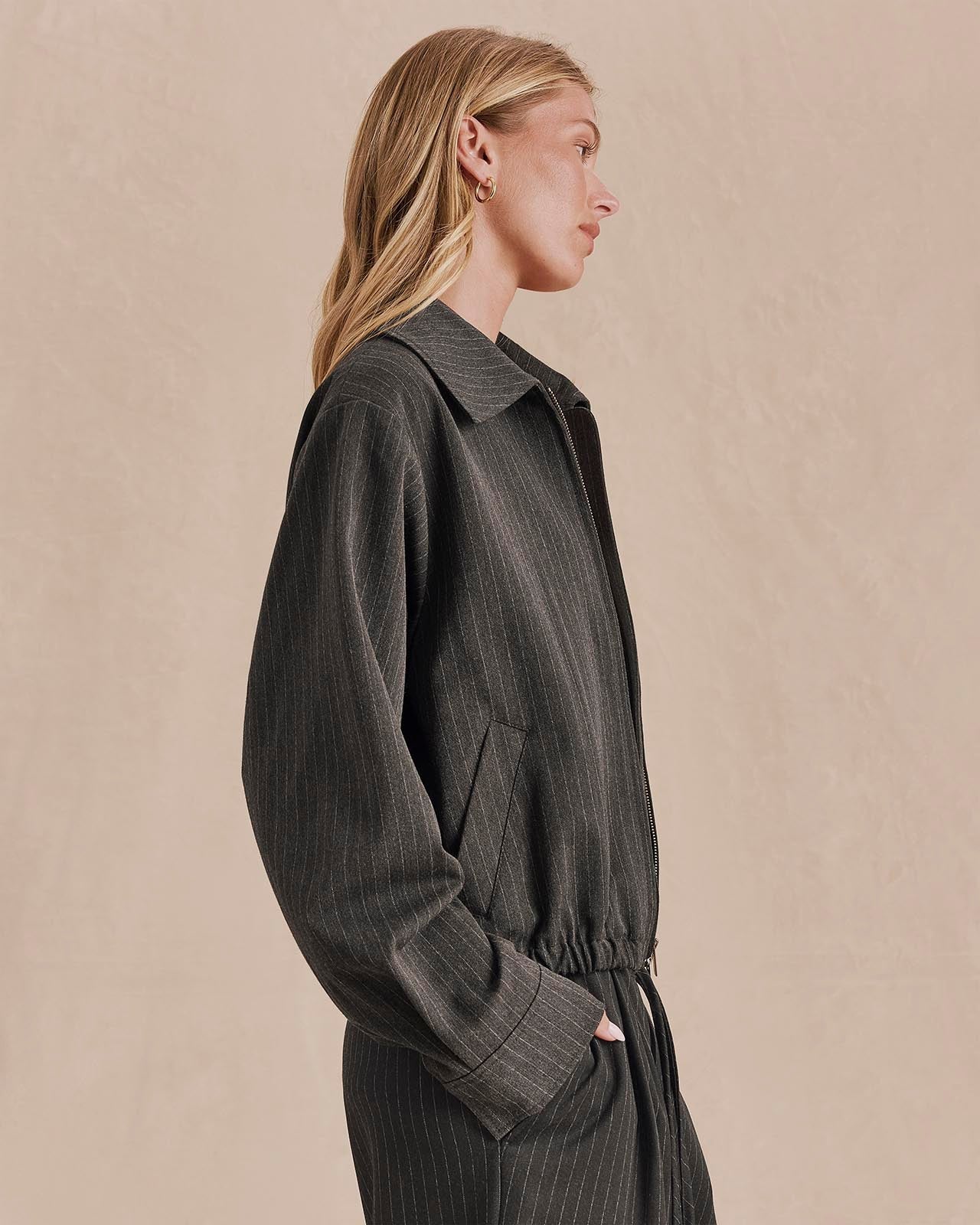 Side view of model wearing charcoal pinstripe zip-up jacket with elastic hem and cuffs