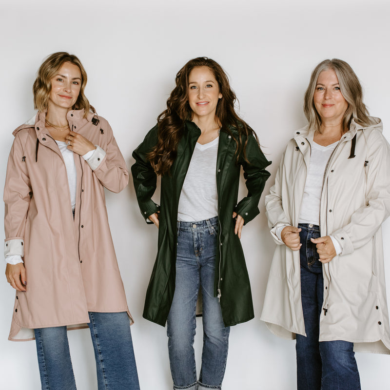 Three women wearing Ilse Jacobsen raincoats in pink, dark green, and cream