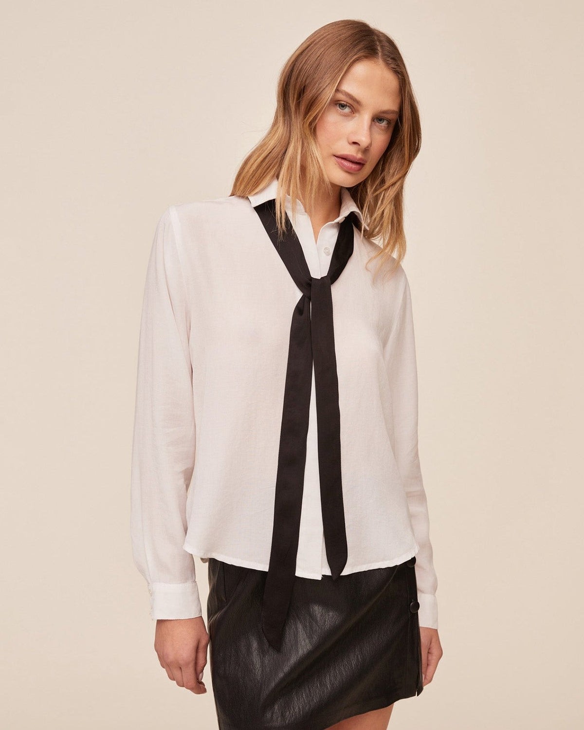 Model wearing white long sleeve shirt with black neck tie and black leather mini skirt