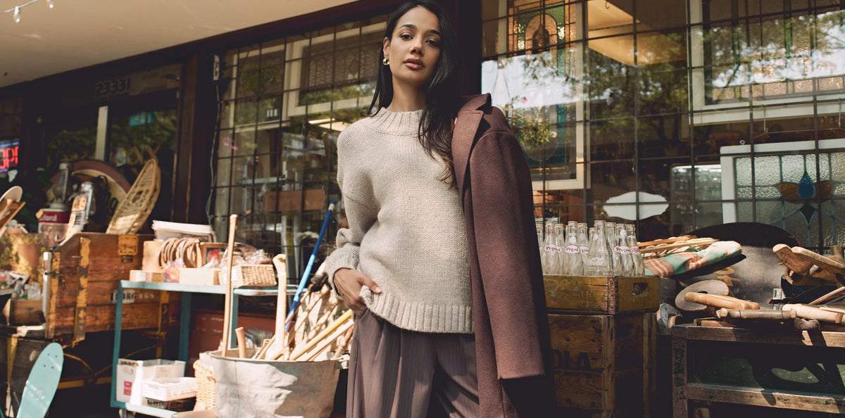 Model wearing cream knit sweater with brown coat draped over shoulder outside vintage shop