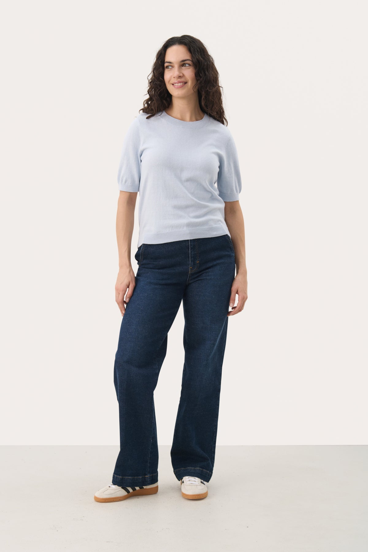 Woman wearing a light gray-blue short-sleeve knit pullover with wide-leg dark jeans and white sneakers.