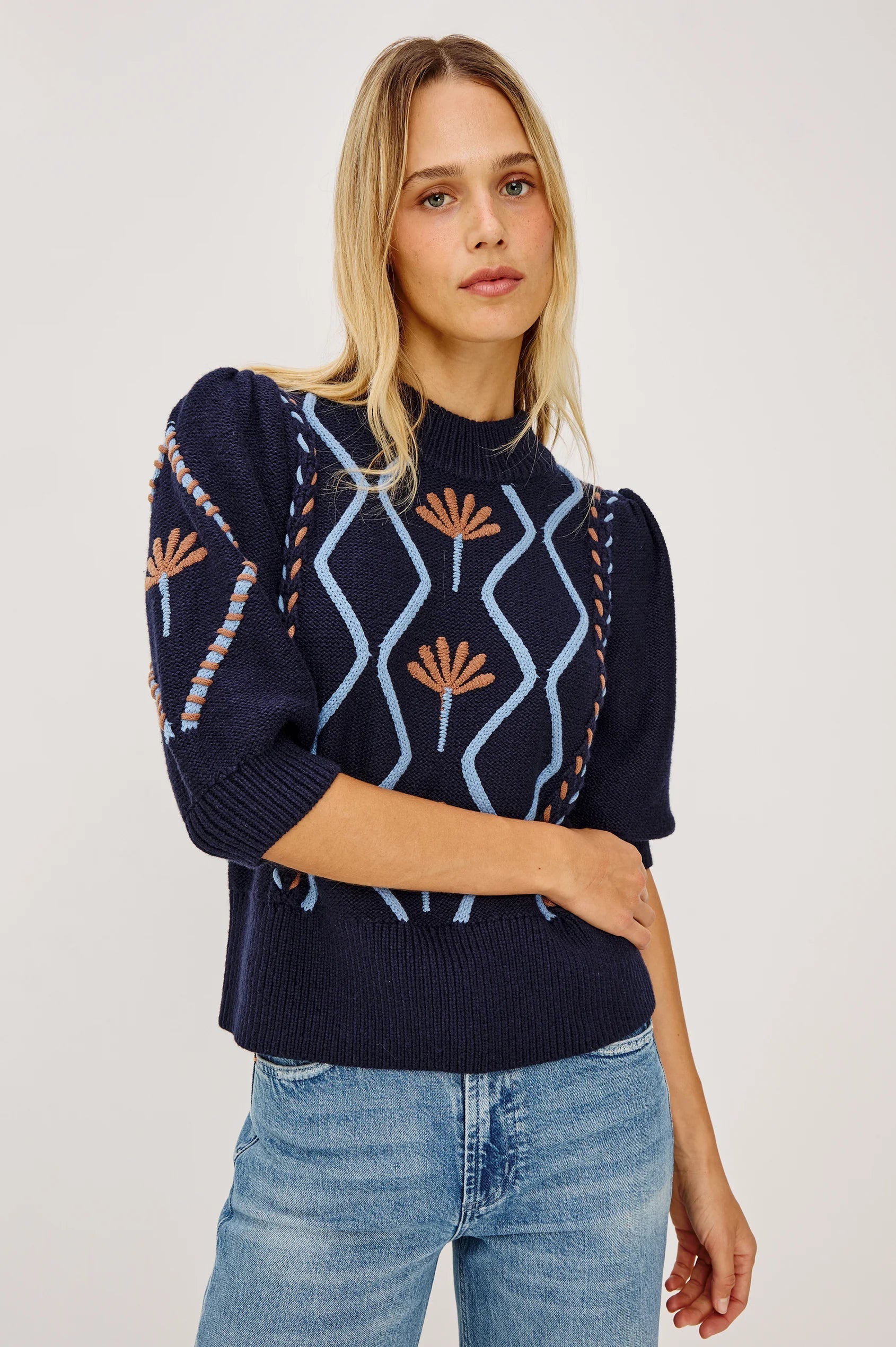Woman wearing navy blue knit sweater with light blue wave cables and terracotta floral embroidery details