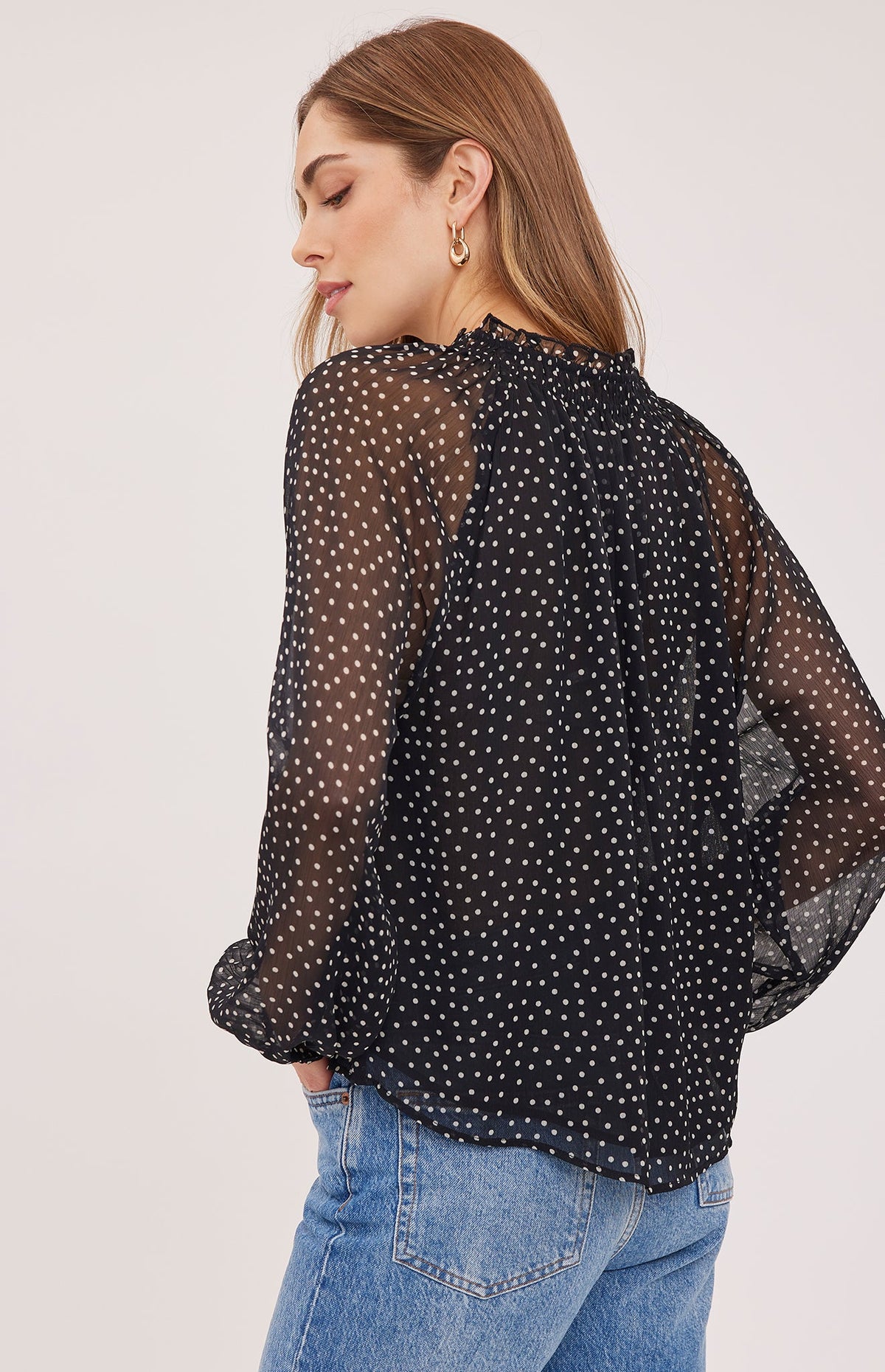 Back view of model wearing black polka dot sheer chiffon blouse with ruffle neckline and long sleeves