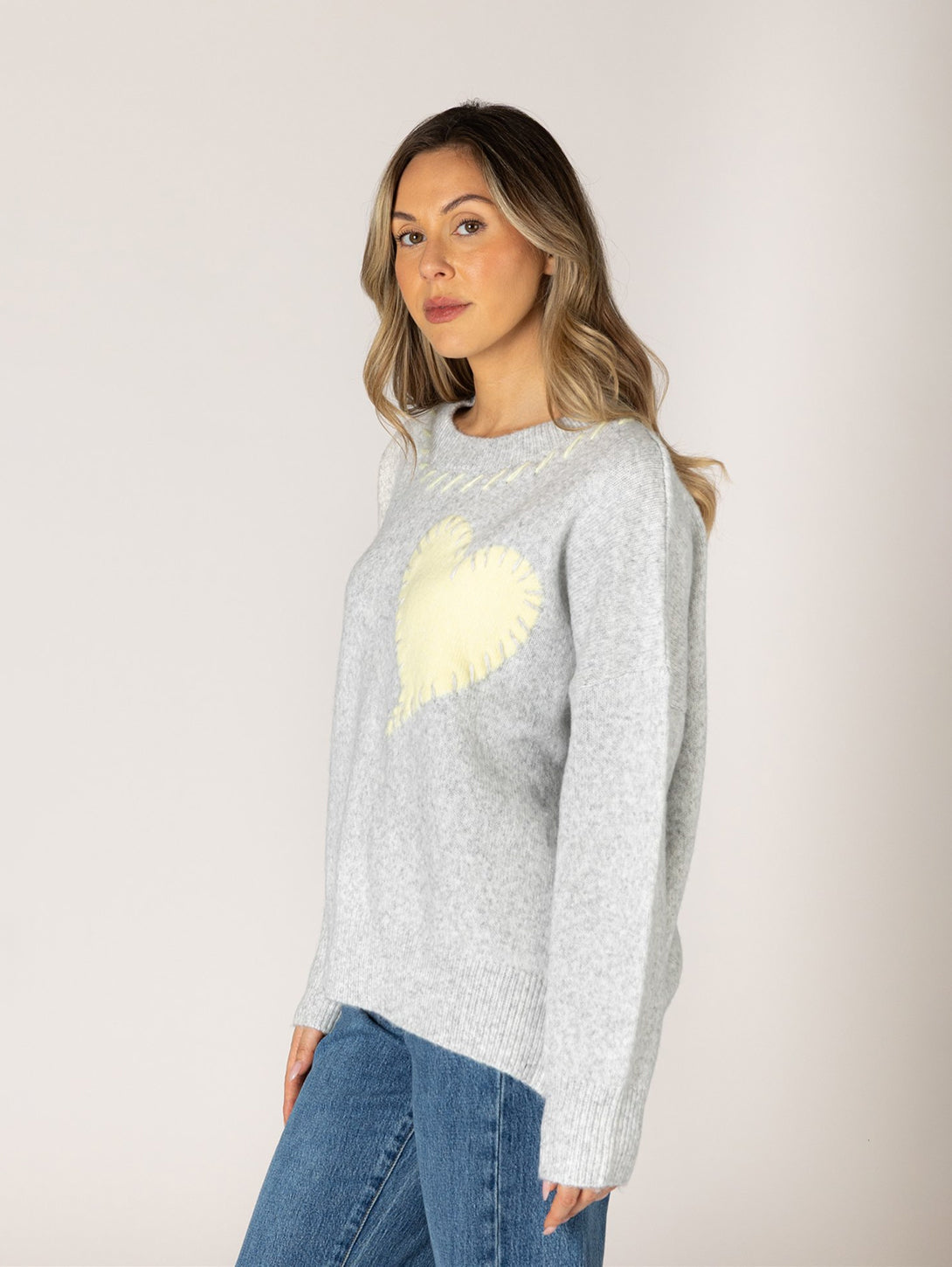 Model wearing grey knit sweater with cream heart motif, side view, paired with blue jeans