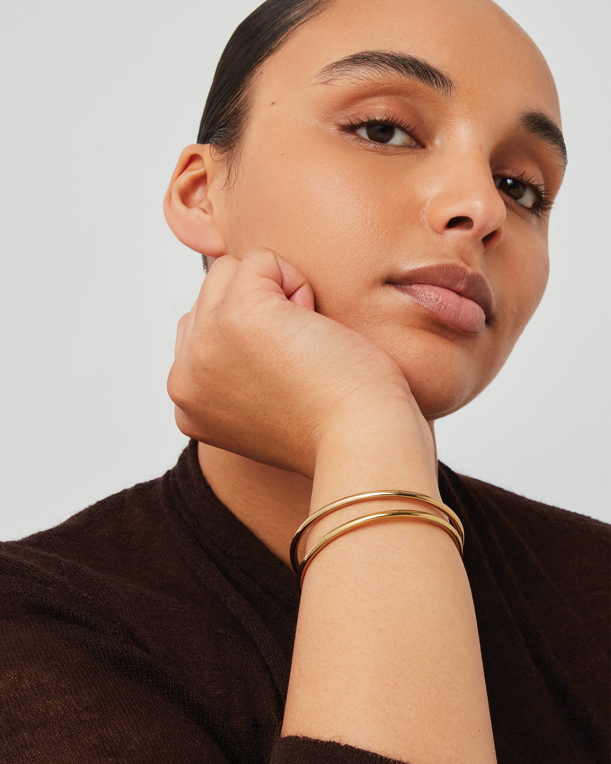 Model wearing two gold bangle bracelets stacked on wrist, resting chin on hand