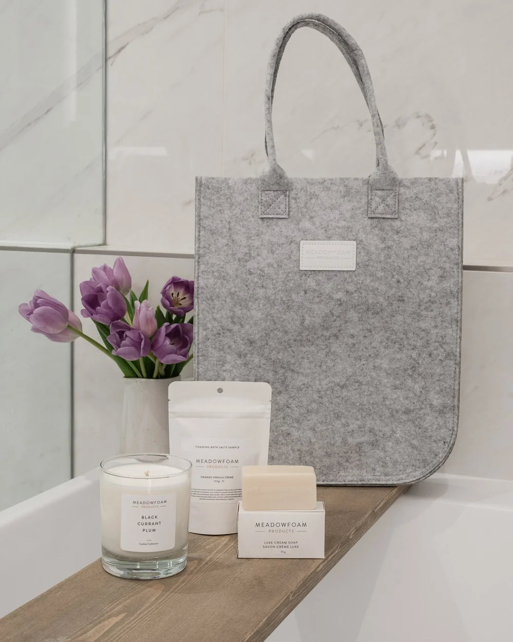 Grey felt tote bag with candle, bath salts, and cream soap beside purple tulips