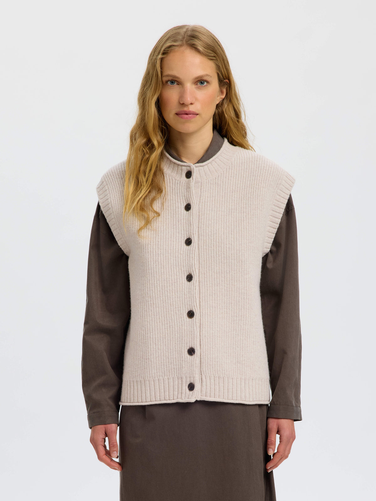 Model wearing a light beige ribbed knit sleeveless vest with dark buttons over brown sleeves