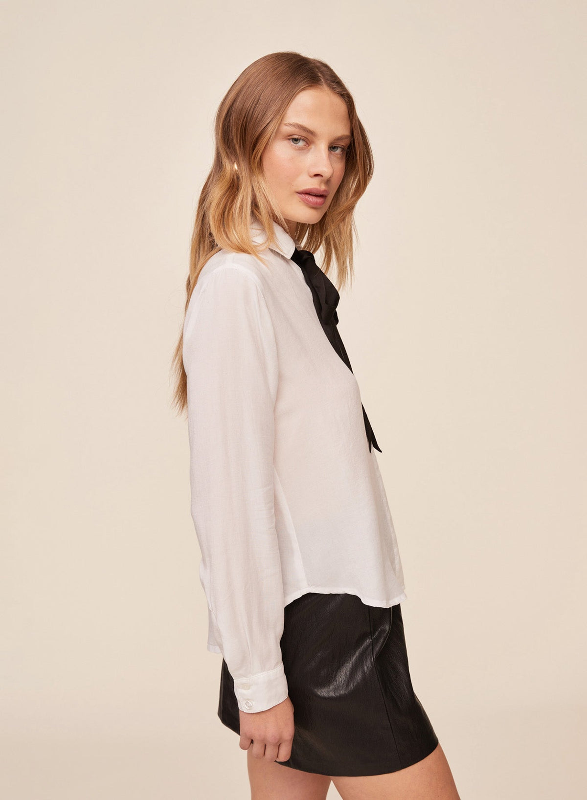 Model wearing white long sleeve shirt with black neck tie and black leather mini skirt, side view