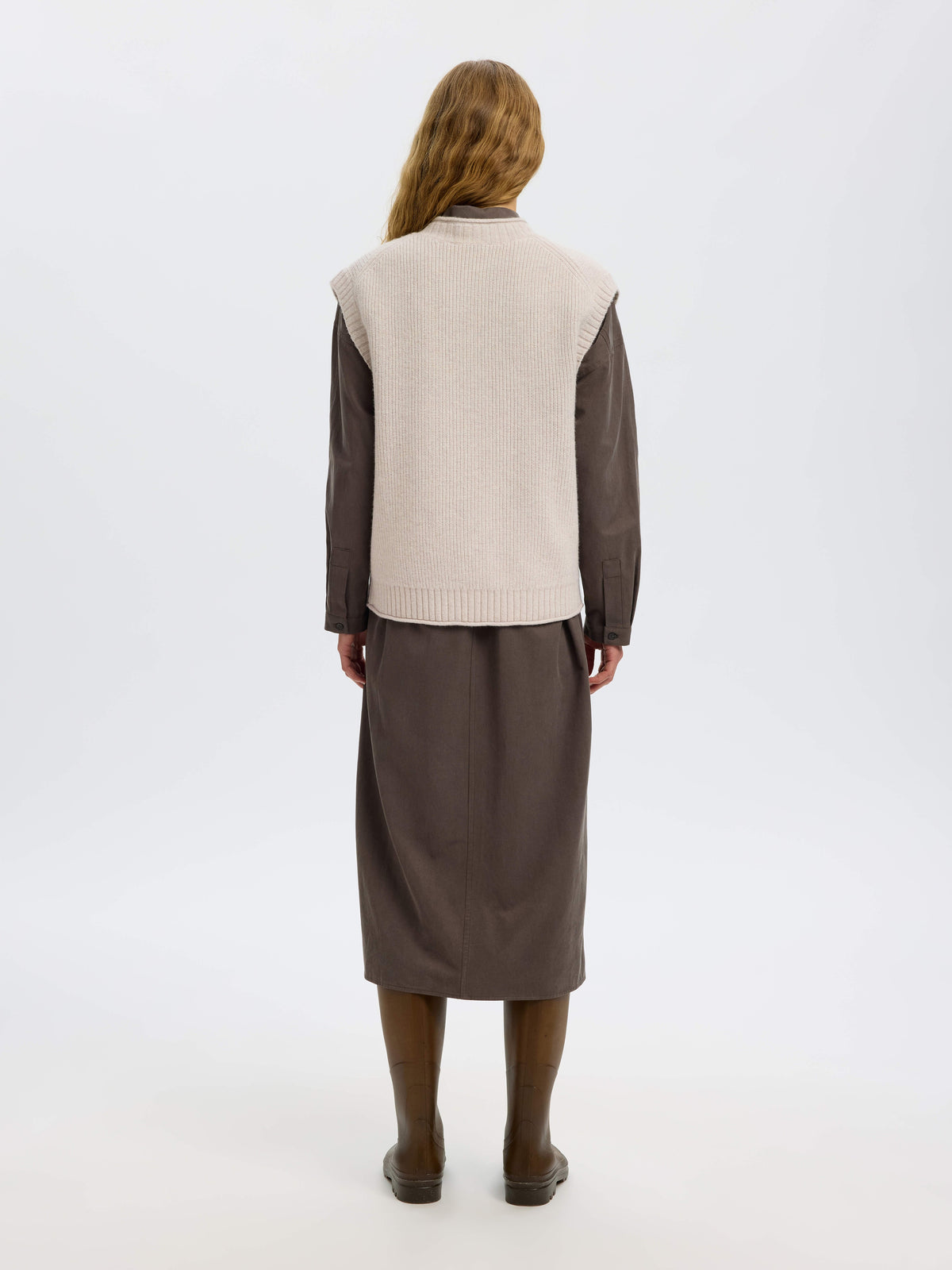 Back view of model wearing cream ribbed knit vest over dark brown shirt and midi skirt.