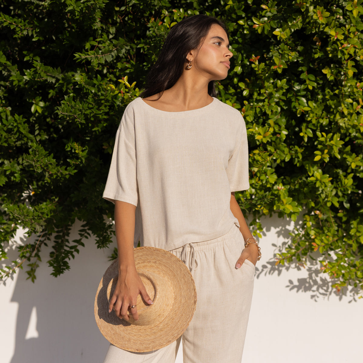 Model wearing cream linen top and matching drawstring pants holding a straw hat outdoors