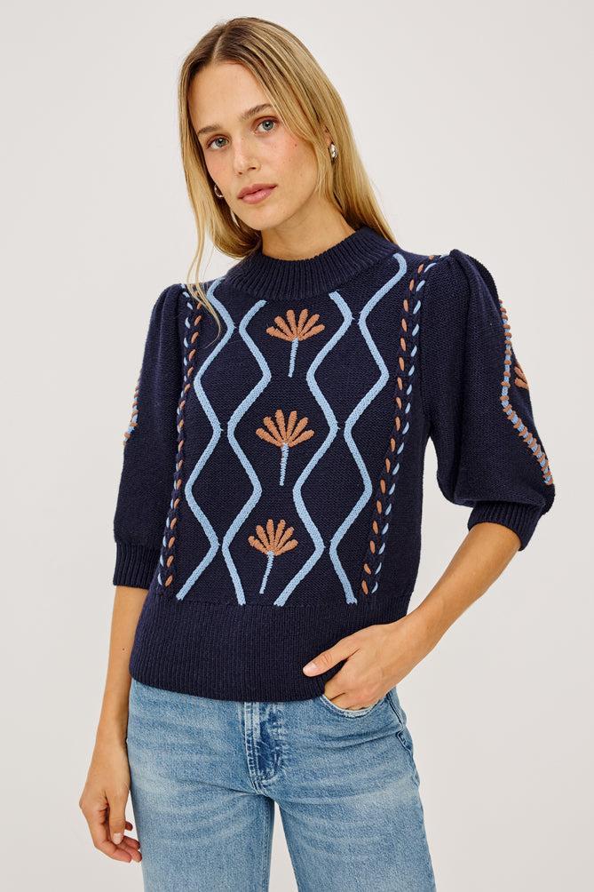 Model wearing navy knit sweater with floral cable pattern and puff sleeves paired with jeans